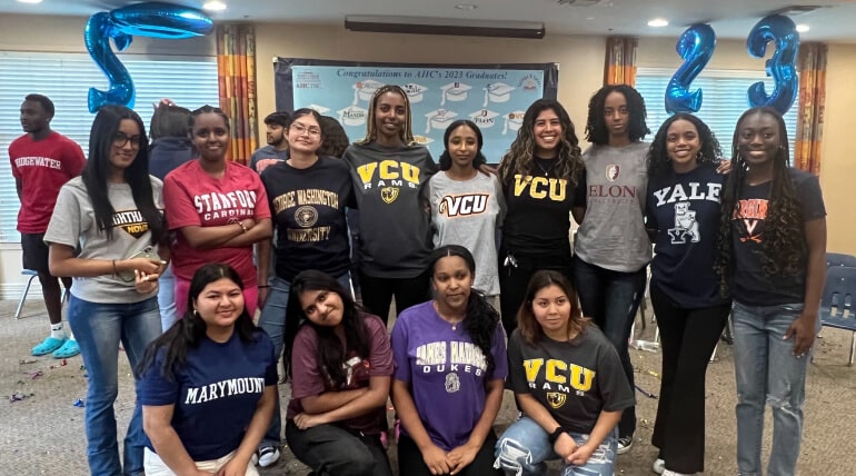 A group of young women pose indoors in college shirts, standing and kneeling in two rows. Blue balloon numbers 23 and a banner celebrate graduation as students declare college decisions on AHC Signing Day.