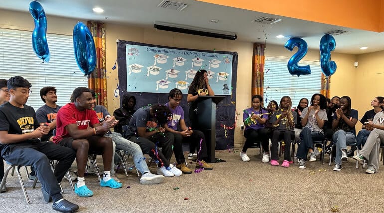 A group of students sits and cheers while a person speaks at a podium. Blue balloons form the numbers 2023 and a congratulatory banner with graduation caps hangs behind them as students declare college decisions on AHC Signing Day. Confetti is scattered on the floor.