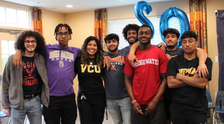 A group of eight smiling young adults stands indoors, arm in arm. Wearing different college t-shirts, they celebrate Students Declare College Decisions on AHC Signing Day with blue balloons forming the number 80 in the background.