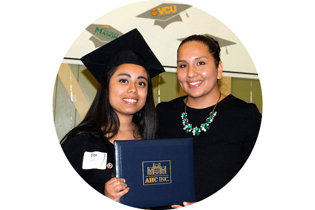 A young woman in a graduation cap and gown holds a diploma folder, standing next to a smiling woman in a black top and green necklace. Graduation cap decorations are visible on the wall behind them.
