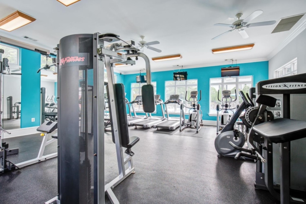 Bright, modern gym with turquoise walls, featuring treadmills, elliptical machines, stationary bikes, weight machines, and large windows letting in natural light. Ceiling fans and TVs are mounted overhead.
