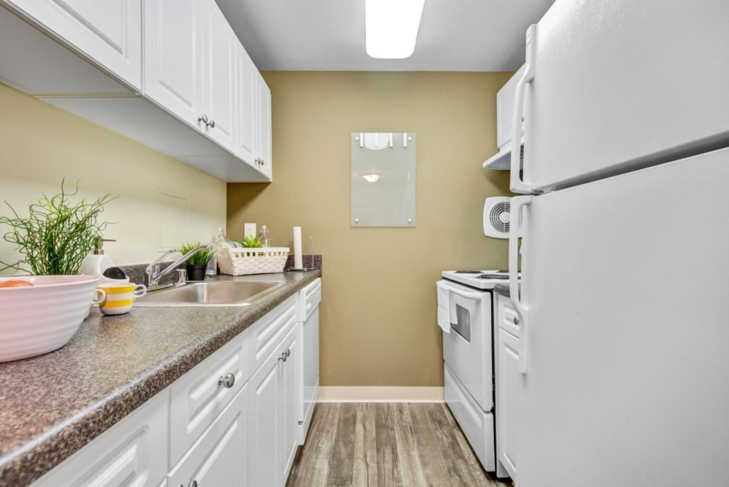 Narrow kitchen with white cabinets, white appliances, brown countertop, and wood-style flooring. A sink, stove, refrigerator, and decorative items like plants and bowls are visible. Walls are painted tan. Bright overhead light.