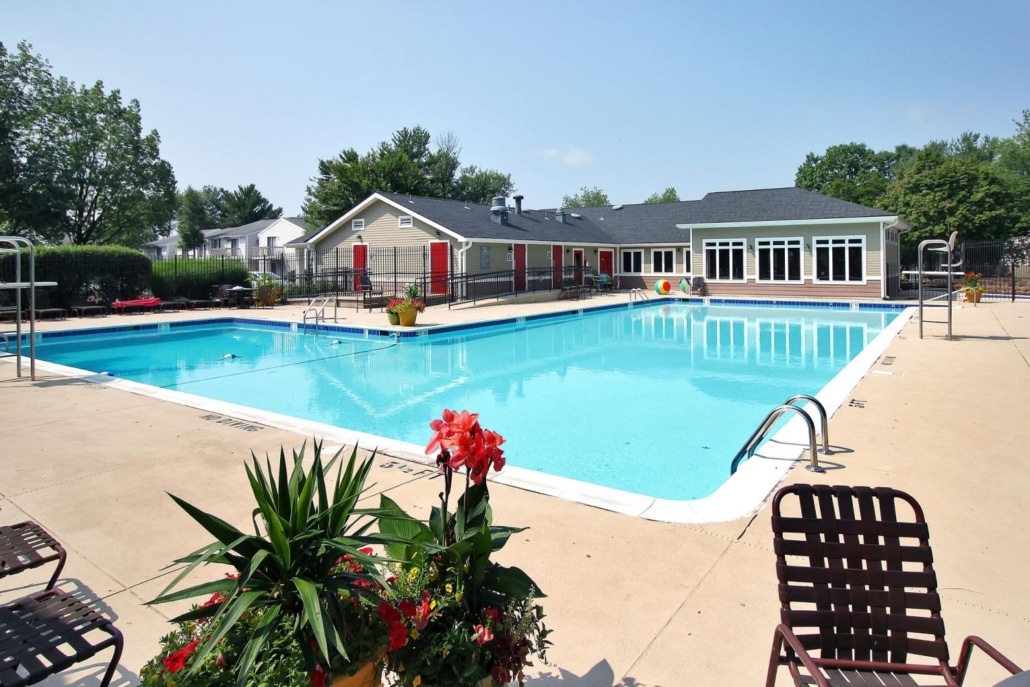 Large outdoor swimming pool with clear blue water, surrounded by a concrete deck, pool chairs, and potted plants. In the background are residential buildings and trees under a clear sky.