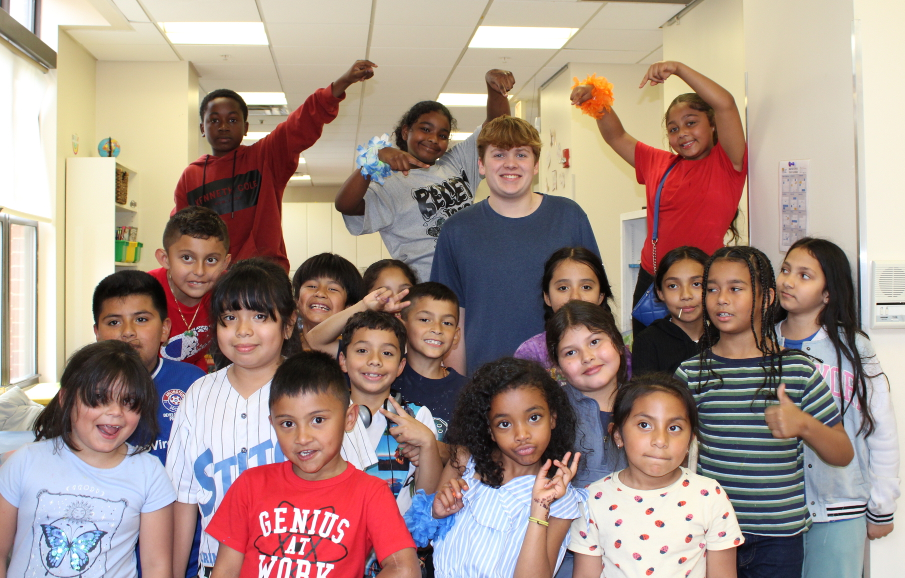A group of children and one adult are smiling and posing together in a classroom setting as AHC celebrates 2025 graduates of the CCR Program. Some kids stand, others sit or kneel, sharing joyful, playful gestures that express excitement.