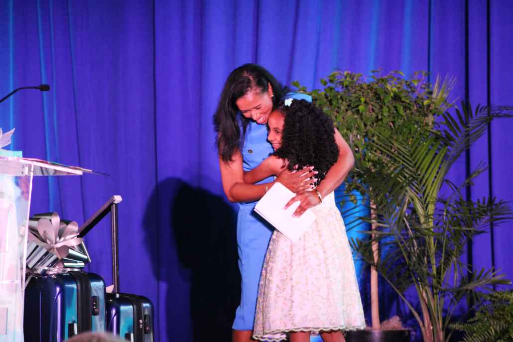 A woman in a blue dress warmly hugs a young girl in a floral dress on stage, beside a podium and suitcases, against a purple backdrop with plants—capturing the joy as AHC launches new website.