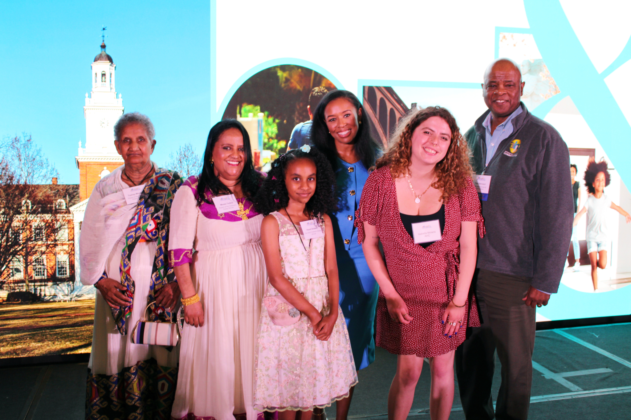 Six people stand together smiling in front of a bright screen displaying a campus scene and abstract designs. The group, in colorful attire, poses for a photo celebrating the AHC Launches New Website event.