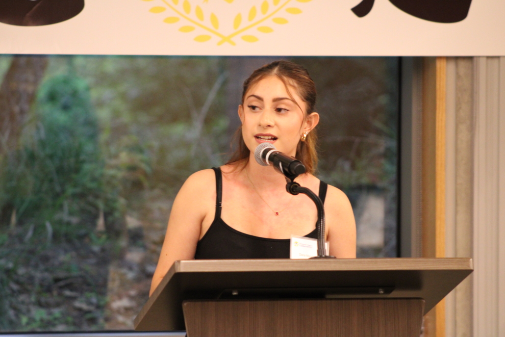 A young woman with light brown hair, wearing a black top, speaks into a microphone at a podium. Behind her, a large window reveals greenery and part of a banner reading A Recipe for Community above.