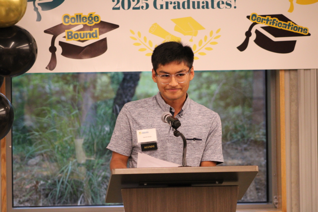 A young man with glasses stands at a podium, smiling, holding papers and speaking into a microphone. Behind him is a banner with graduation caps and words like 2025 Graduates, College Bound, Certifications—a recipe for community success.