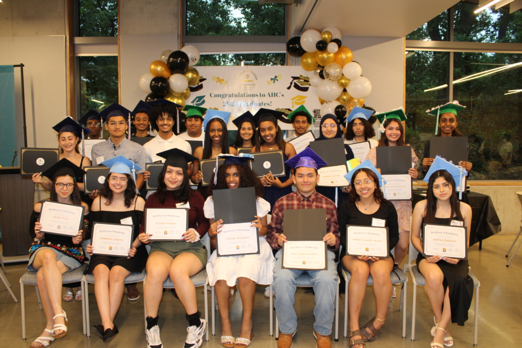 A group of students in graduation caps and gowns pose indoors, holding certificates and folders. Smiling in two rows with balloons and a Congratulations banner behind them, they embody a recipe for community and shared achievement.