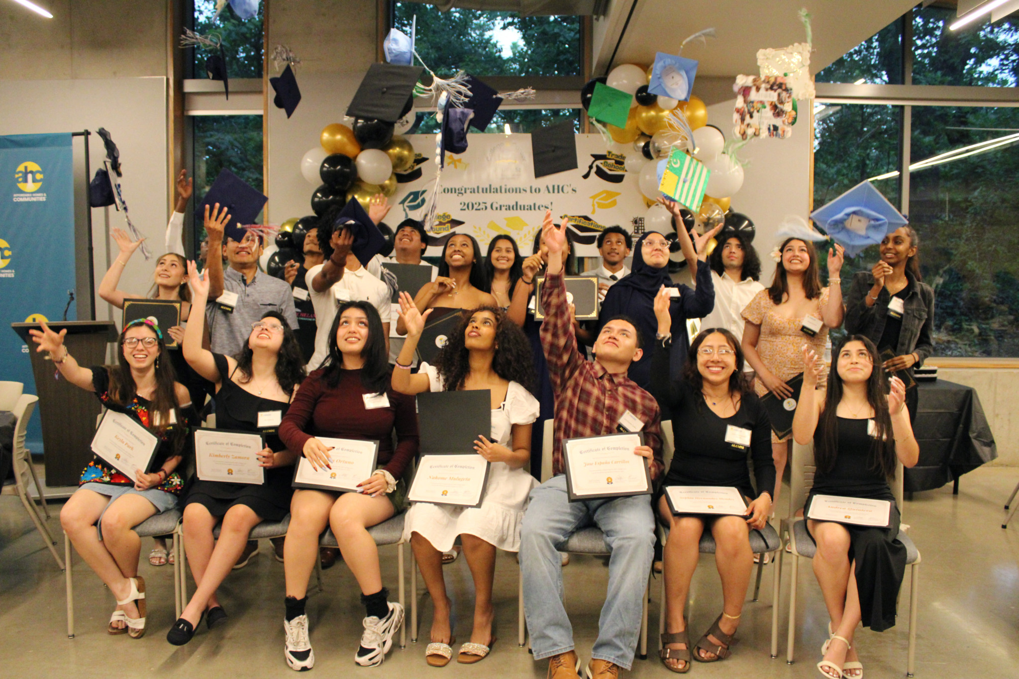 A group of graduates, dressed in semi-formal attire, celebrate A Recipe for Community by tossing their caps and holding certificates in a decorated room filled with congratulatory banners and balloons.