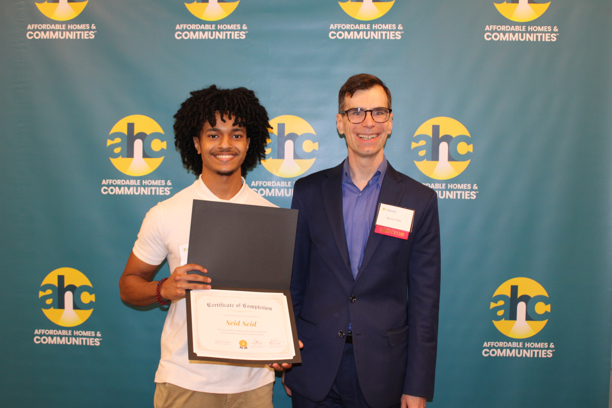 Two people stand in front of an Affordable Homes & Communities backdrop. One holds a certificate of completion and smiles, while the other, wearing glasses and a suit, stands beside him as AHC celebrates 2025 graduates of the CCR Program.