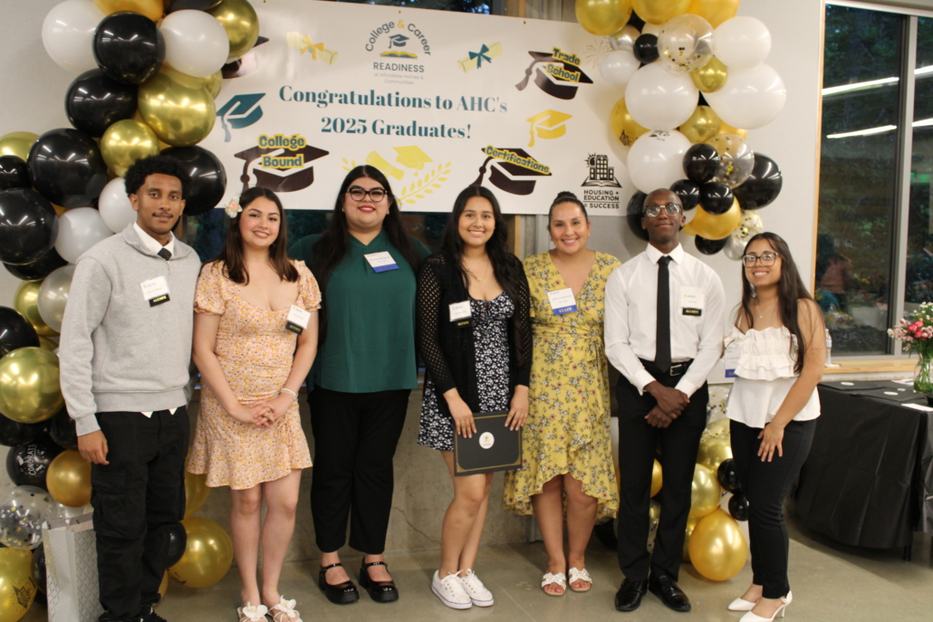 Seven students dressed in semi-formal attire stand smiling in front of a decorated backdrop with balloons and a sign that reads “Congratulations to AHC’s 2023 Graduates! College Bound”—a true recipe for community success.