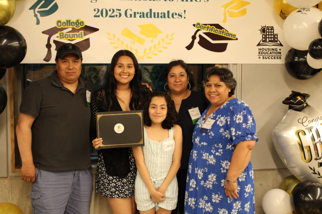 A young woman holds a diploma and stands with her family in front of a congratulatory banner for 2025 graduates. Smiling and surrounded by balloons, they celebrate together—a true recipe for community at this special event.