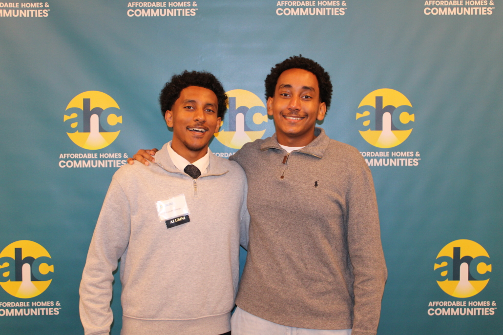Two men smiling and posing together in front of a blue backdrop displaying the Affordable Homes & Communities logo, embodying A Recipe for Community. One wears a light gray sweater and collared shirt; the other wears a gray pullover.