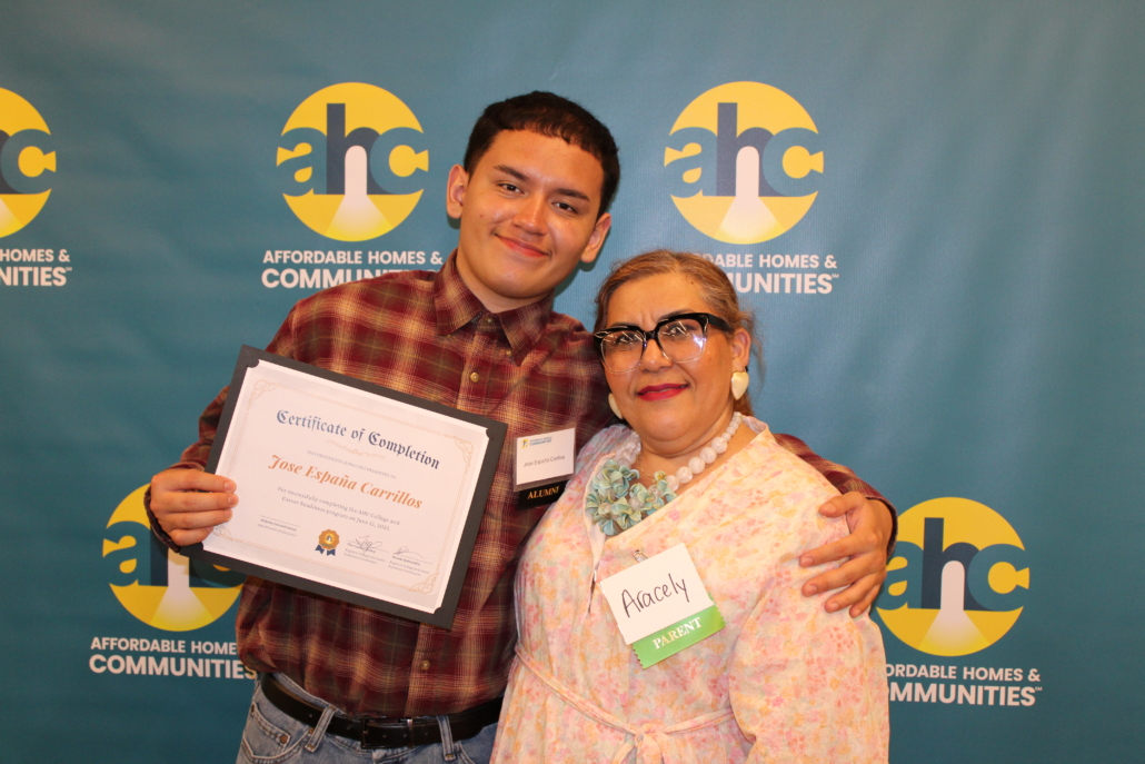 A young man holds a certificate of completion and stands next to an older woman named Anacely. They are smiling in front of an Affordable Homes & Communities backdrop, celebrating their part in A Recipe for Community.