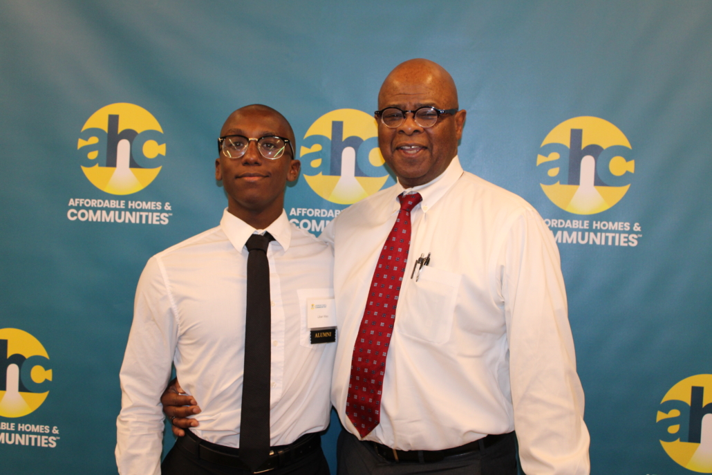 Two men stand smiling together in front of a blue backdrop with “AHC Affordable Homes & Communities” logos, embodying A Recipe for Community. Both wear glasses, white shirts, and ties; one man has his arm around the other.