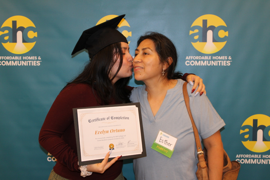 A graduate in a cap kisses a woman, likely his mother, on the cheek while holding a certificate. The smiling woman, Esther Parent, wears a nametag. Behind them is the Affordable Homes & Communities logo and A Recipe for Community message.