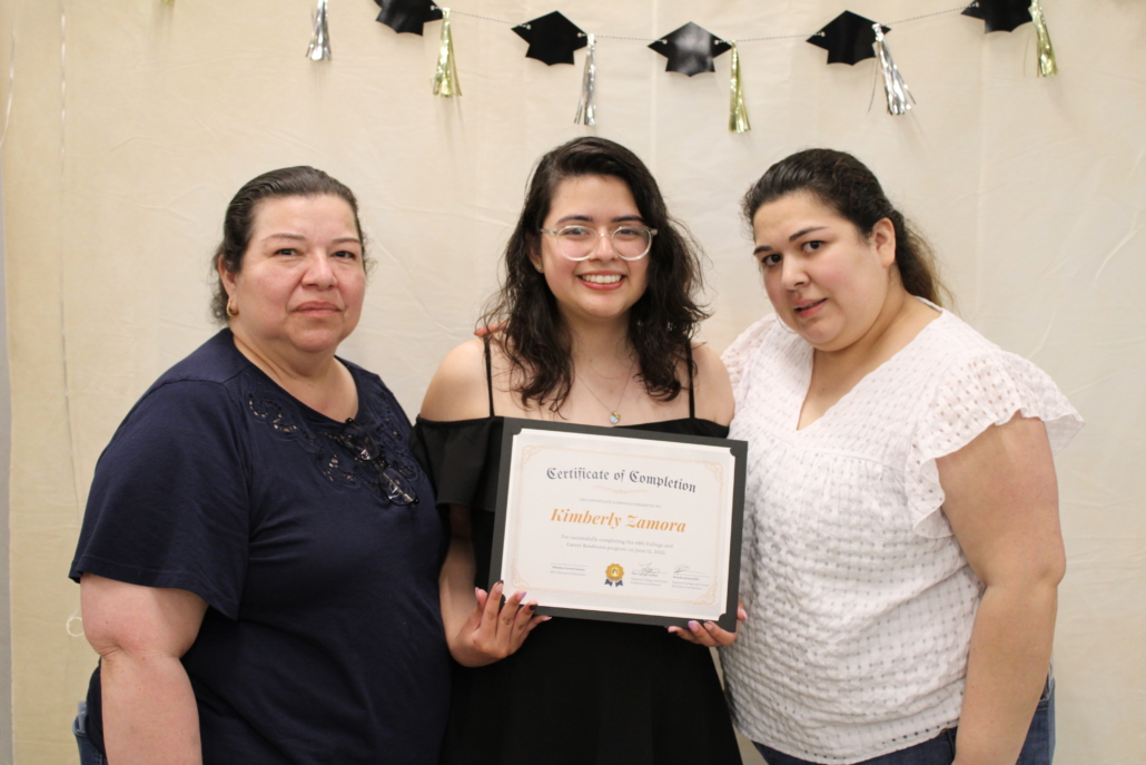 Three women stand together, smiling, in front of a graduation-themed backdrop. The woman in the center holds a Certificate of Completion. A Recipe for Community shines through as her friends on either side support her, all dressed in casual clothing.