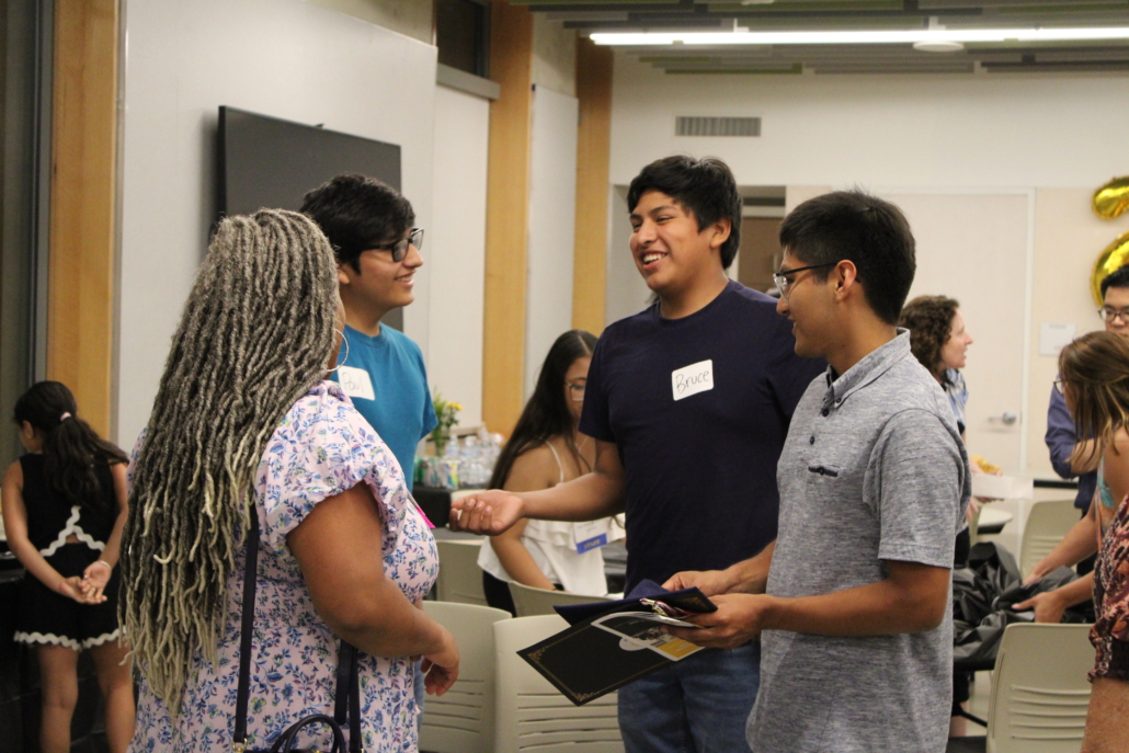 A group of people stand and converse in a brightly lit room. One woman with long, gray braids faces three smiling men, one holding a folder—a true recipe for community. Additional people are seated and mingling in the background.