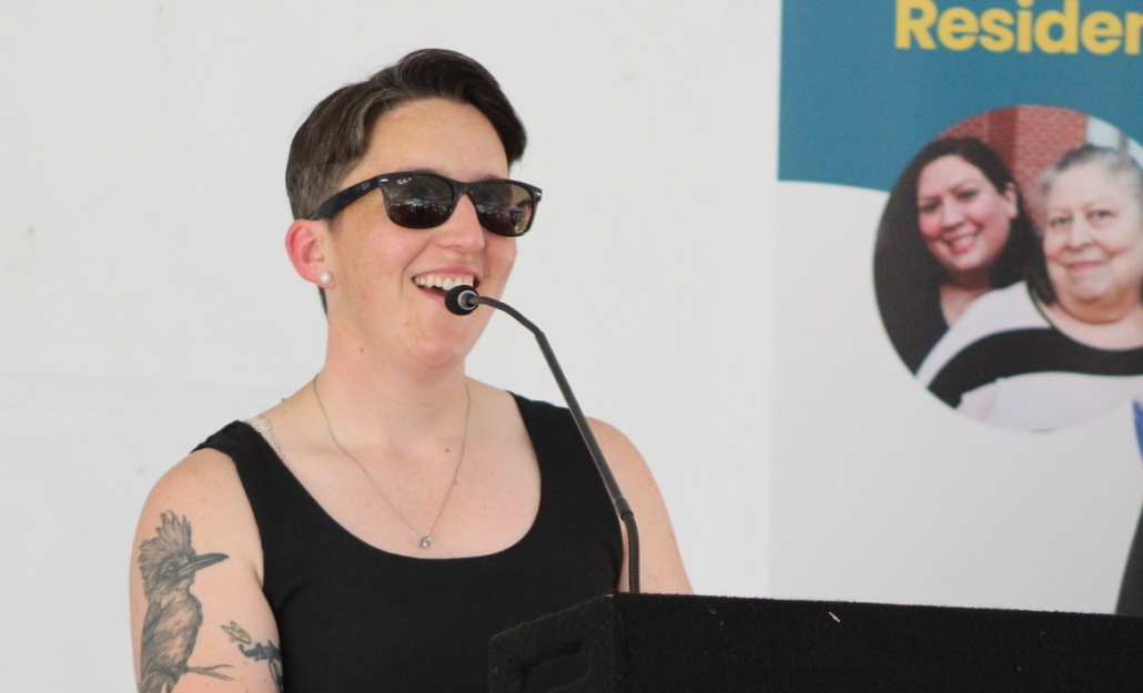 Person wearing sunglasses and a black tank top speaks at the podium, smiling, during the Allium Place affordable housing ribbon-cutting. A bird tattoo is visible on their left arm, with a poster of two women in the background.