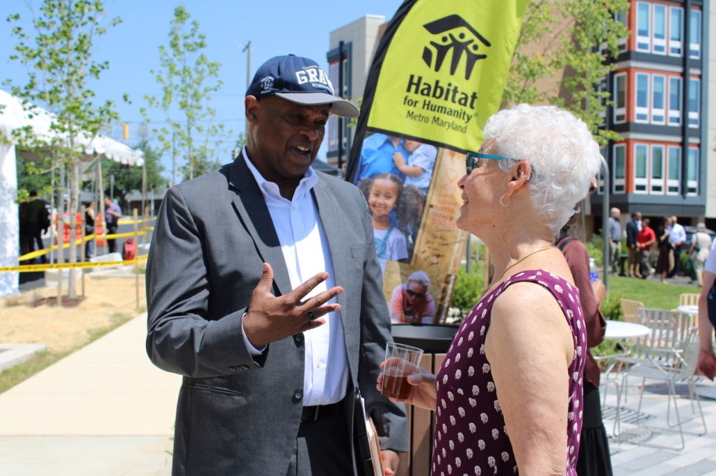 A man in a gray suit and cap talks with a woman in a sleeveless polka-dot top at the Allium Place affordable housing ribbon-cutting, an outdoor Habitat for Humanity event; a banner and people are visible in the background.