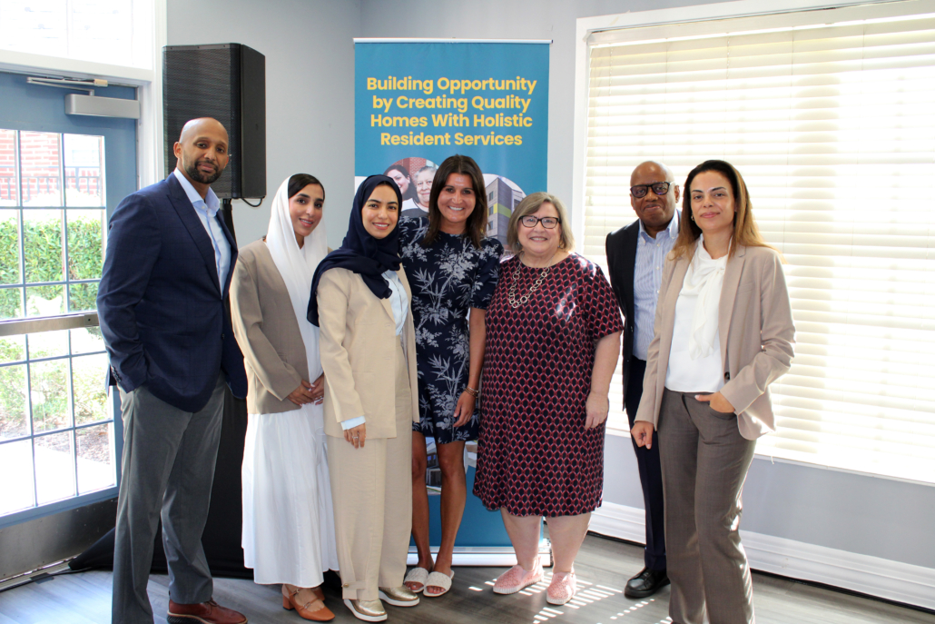 A group of seven people, dressed in business and semi-formal attire, stand smiling together indoors in front of a banner about building quality homes as AHC engages the Georgetown EMBA team to explore a scalable resident services model.