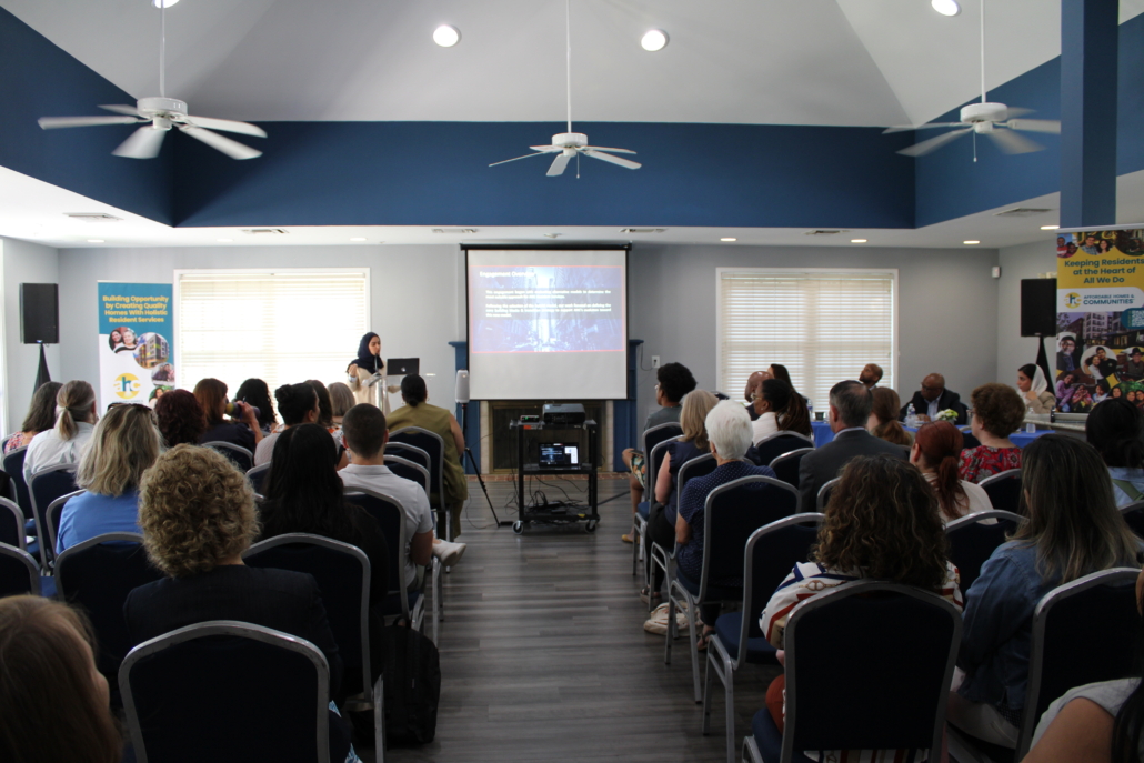 A woman speaks at the front of a room filled with seated people, presenting a slideshow on how AHC Engages Georgetown EMBA Team to Explore Scalable Resident Services Model. The blue-walled room features high ceilings, ceiling fans, and sunlit windows.