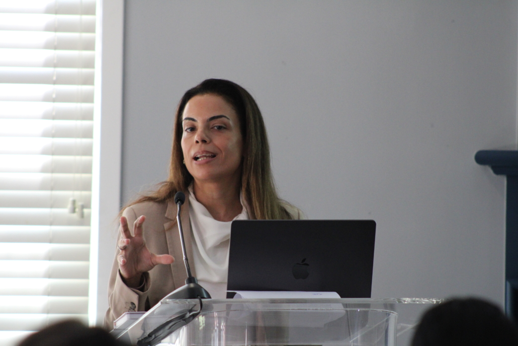 A woman stands behind a clear podium, speaking and gesturing with her hand. She is in front of an open laptop, discussing how AHC engages Georgetown EMBA Team to explore a scalable resident services model, with window blinds and a microphone visible nearby.