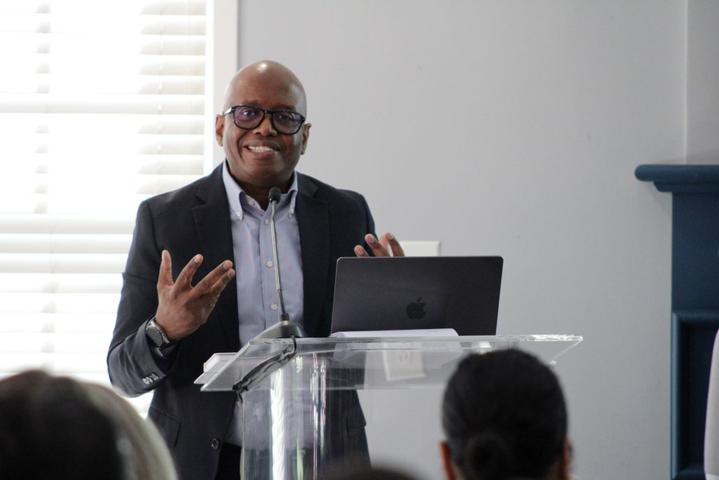 A man in a suit and glasses smiles while speaking at a podium with a laptop, gesturing with one hand, as he discusses how AHC engages the Georgetown EMBA team to explore a scalable resident services model. An audience listens attentively; blinds cover the window behind him.