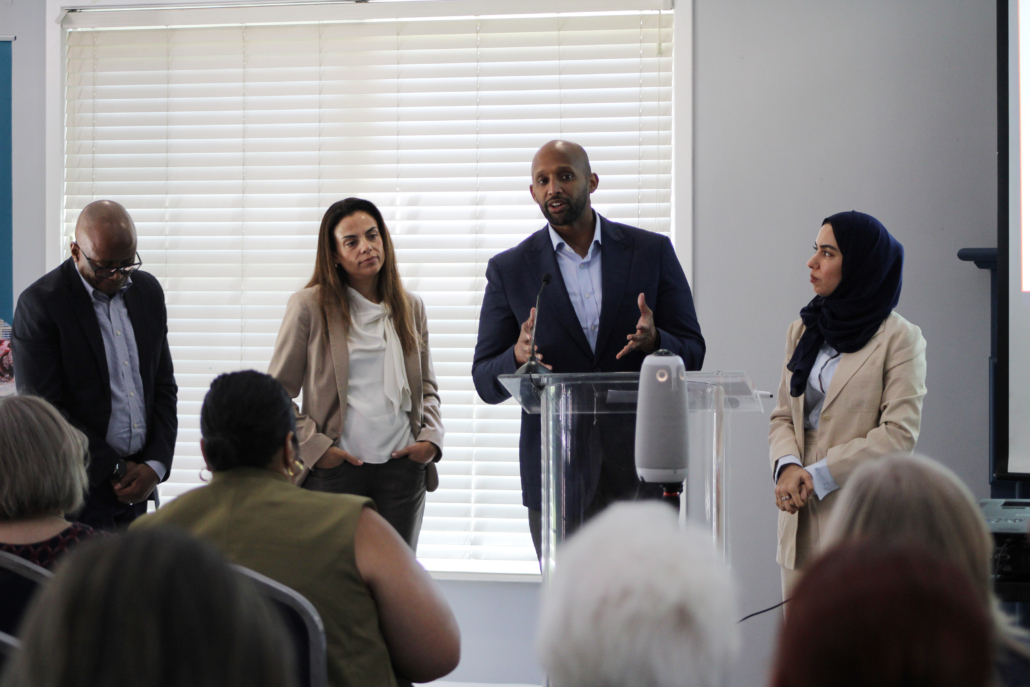 Four people stand at the front of a room, three facing an audience and one speaking at a clear podium, as AHC engages the Georgetown EMBA team to explore a scalable resident services model; blinds cover the window behind them.