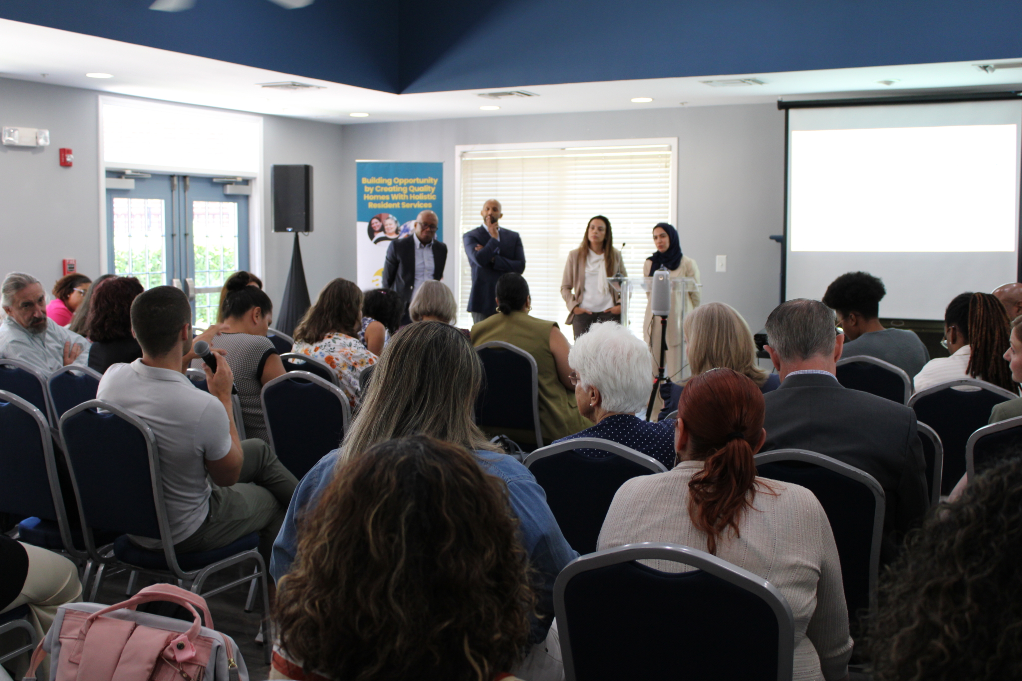 A diverse group of people faces a panel of four speakers at the front of a meeting room, as AHC engages the Georgetown EMBA Team to explore a scalable resident services model. Attendees listen attentively and take notes, with a blank projection screen and banner in view.