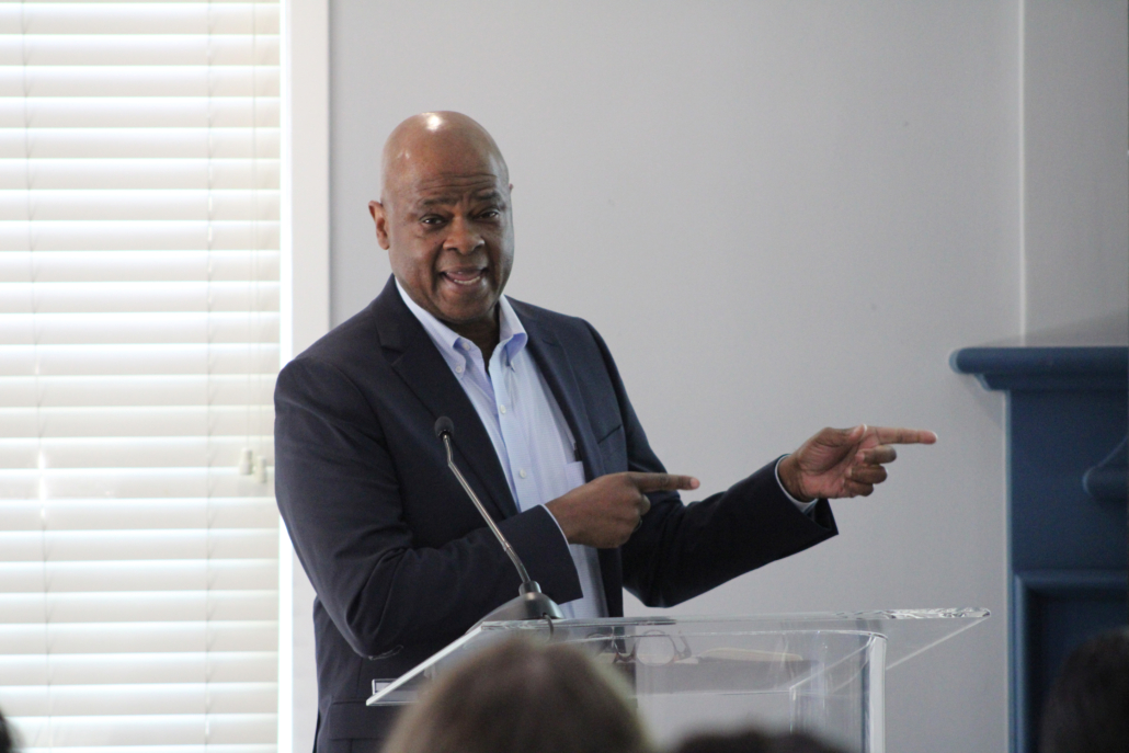 A man in a suit stands at a clear podium, addressing an audience as he discusses AHC Engages Georgetown EMBA Team to Explore Scalable Resident Services Model in a well-lit room with white blinds, gesturing with both hands to his right.