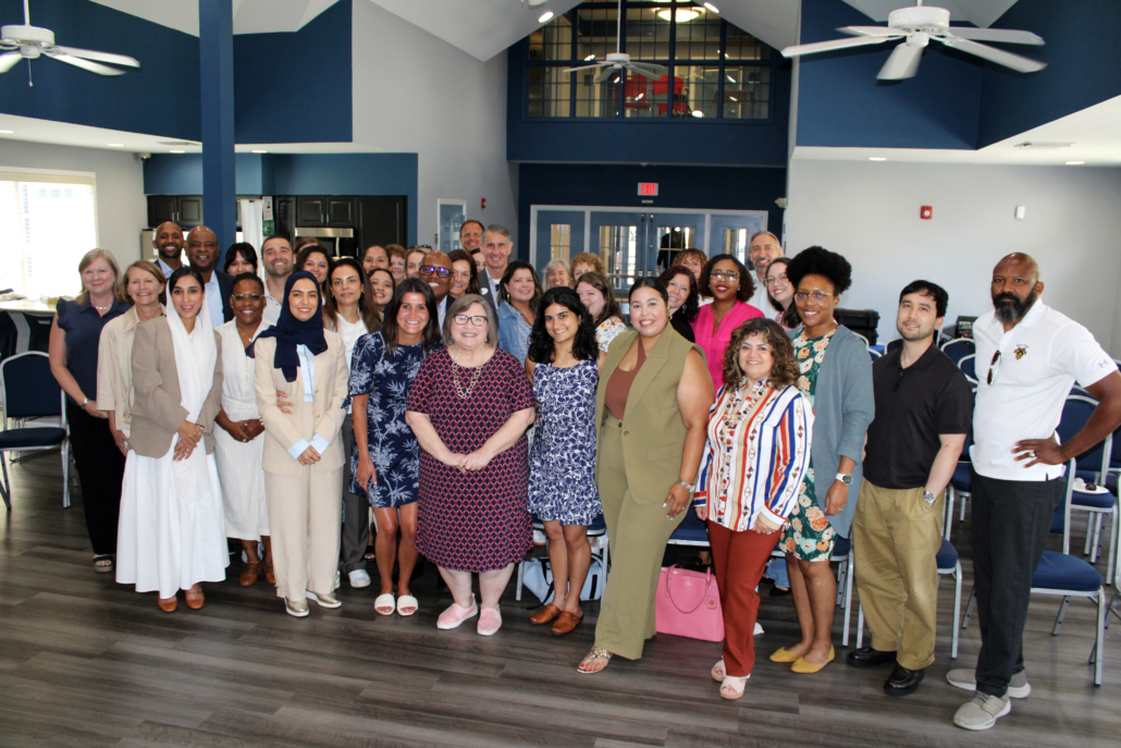 A diverse group of people, both men and women, pose and smile together in a spacious, well-lit room as AHC Engages Georgetown EMBA Team to Explore Scalable Resident Services Model. Blue and white walls, high ceilings, and empty chairs complete the scene.