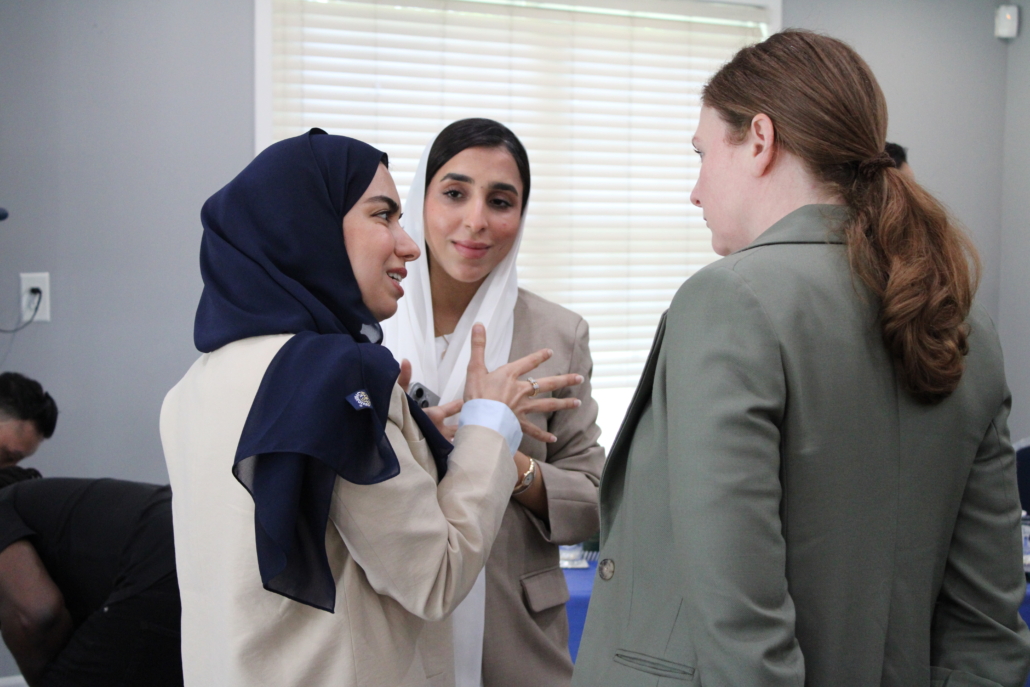 Three women in business attire stand together indoors, engaged in a conversation about how AHC engages Georgetown EMBA Team to explore a scalable resident services model. Two wear hijabs and light blazers; the third, with long hair tied back, sports a green blazer.