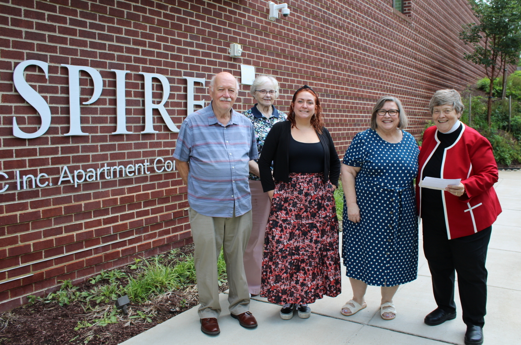 Five people stand smiling outside a brick building next to a sign that reads SPIRE, celebrating the New Period Pantry Opens at Gates of Ballston. The group is casually dressed, with greenery and a sidewalk in the background.