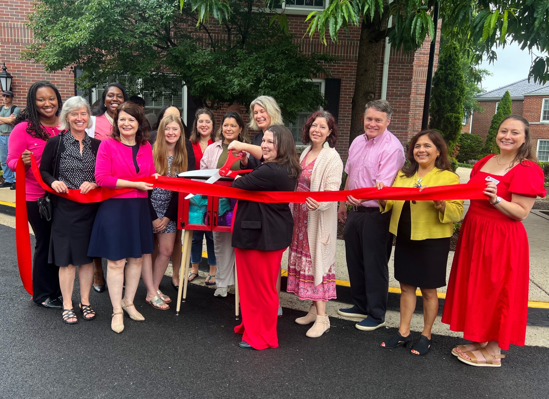 A group of people smiling and standing outside in front of a building, holding a long red ribbon. One woman in the center cuts the ribbon with large scissors as the New Period Pantry opens at Gates of Ballston during a celebratory event.
