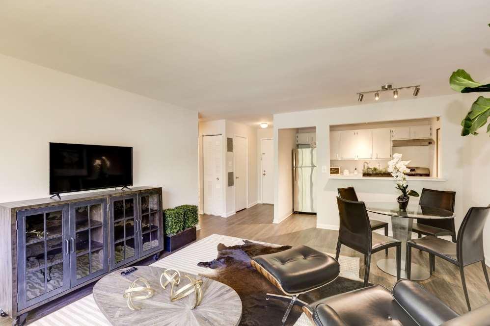 Modern living and dining area with a glass dining table, black chairs, TV on a console, cowhide rug, and a view into a white kitchen with stainless steel appliances and bar counter. Bright, neutral decor.