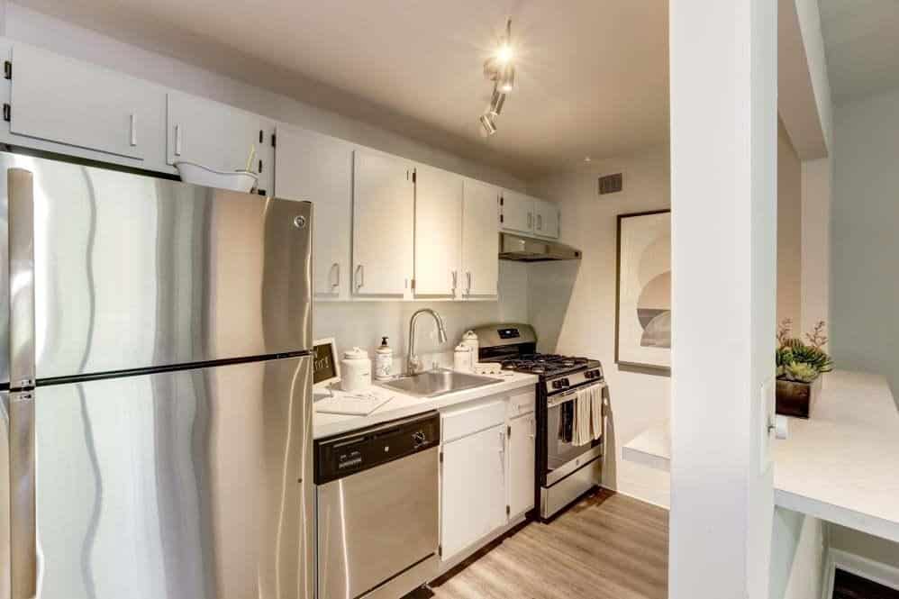 Modern kitchen with stainless steel refrigerator, dishwasher, oven, and white cabinets. A small sink and countertop are visible, along with a potted plant and abstract wall art in the background.