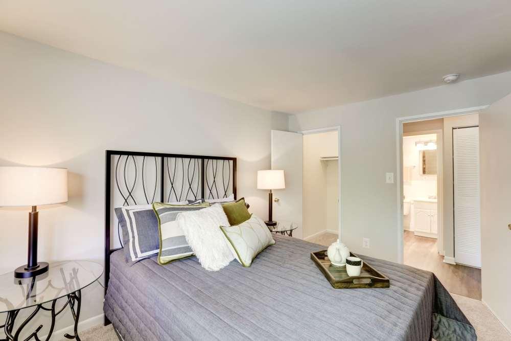 A cozy bedroom with a neatly made bed featuring gray and white pillows, two bedside tables with lamps, a tray on the bed, an open closet, and a view into a brightly lit bathroom.