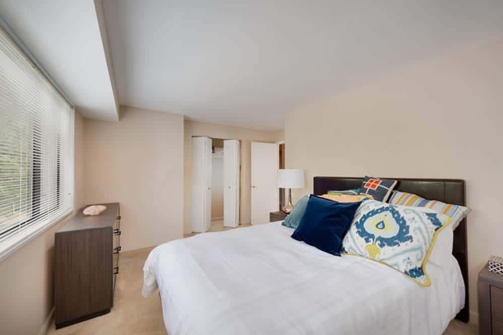 A bright bedroom with a large window, a bed with colorful pillows, a nightstand with a lamp, and a dresser. The walls are light-colored and there is a closet with white doors in the background.