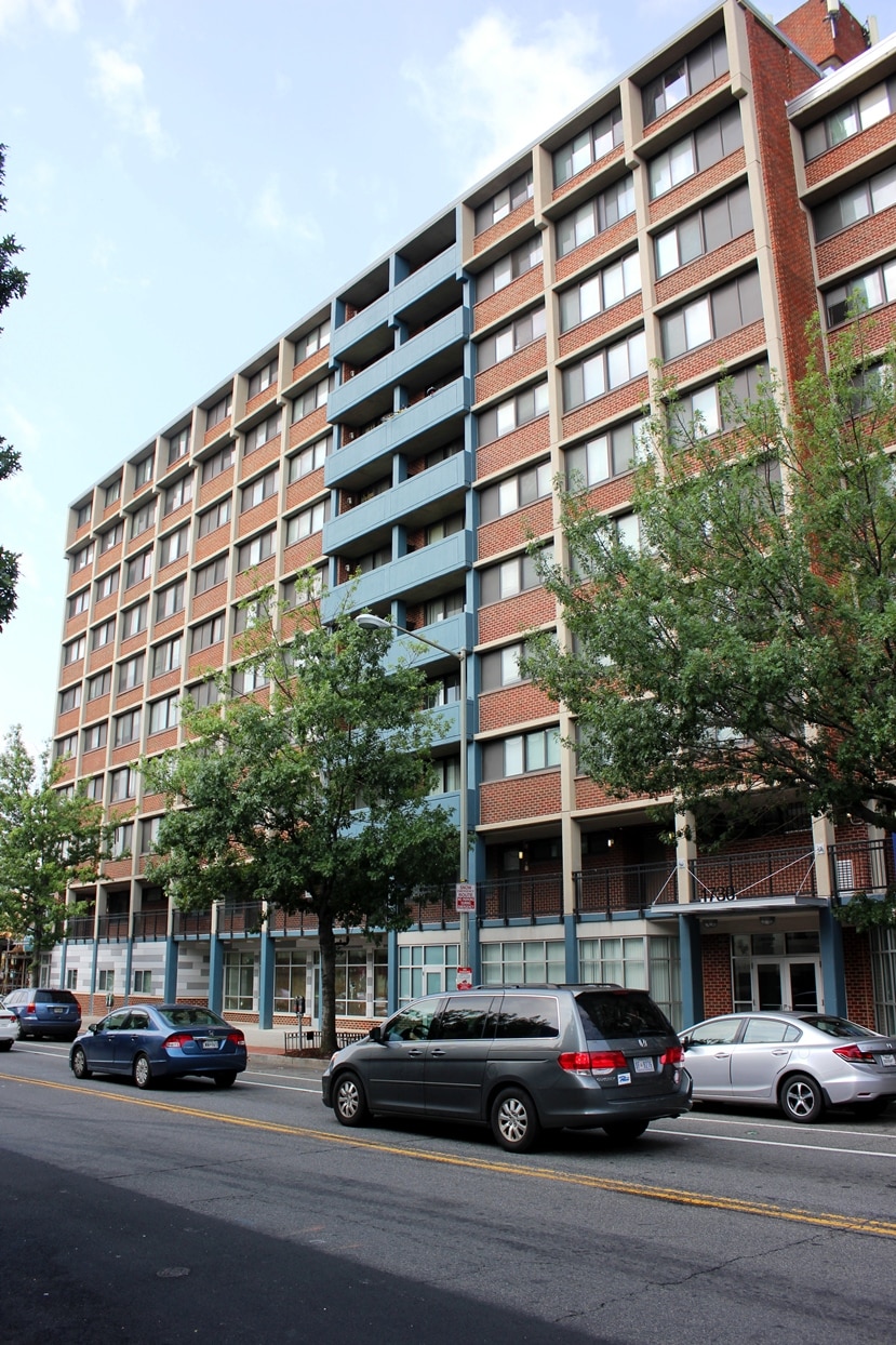 A multi-story brick apartment building with blue accents, large windows, and balconies, with several trees and parked cars lining the street in front.