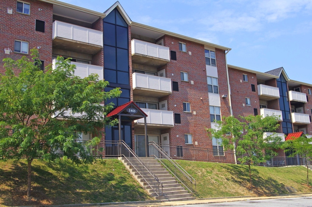 Three-story brick apartment building with balconies, large central windows, and red awnings above entrances. Steps lead up to the main door. Trees and grass line the sidewalk in front. Blue sky in the background.