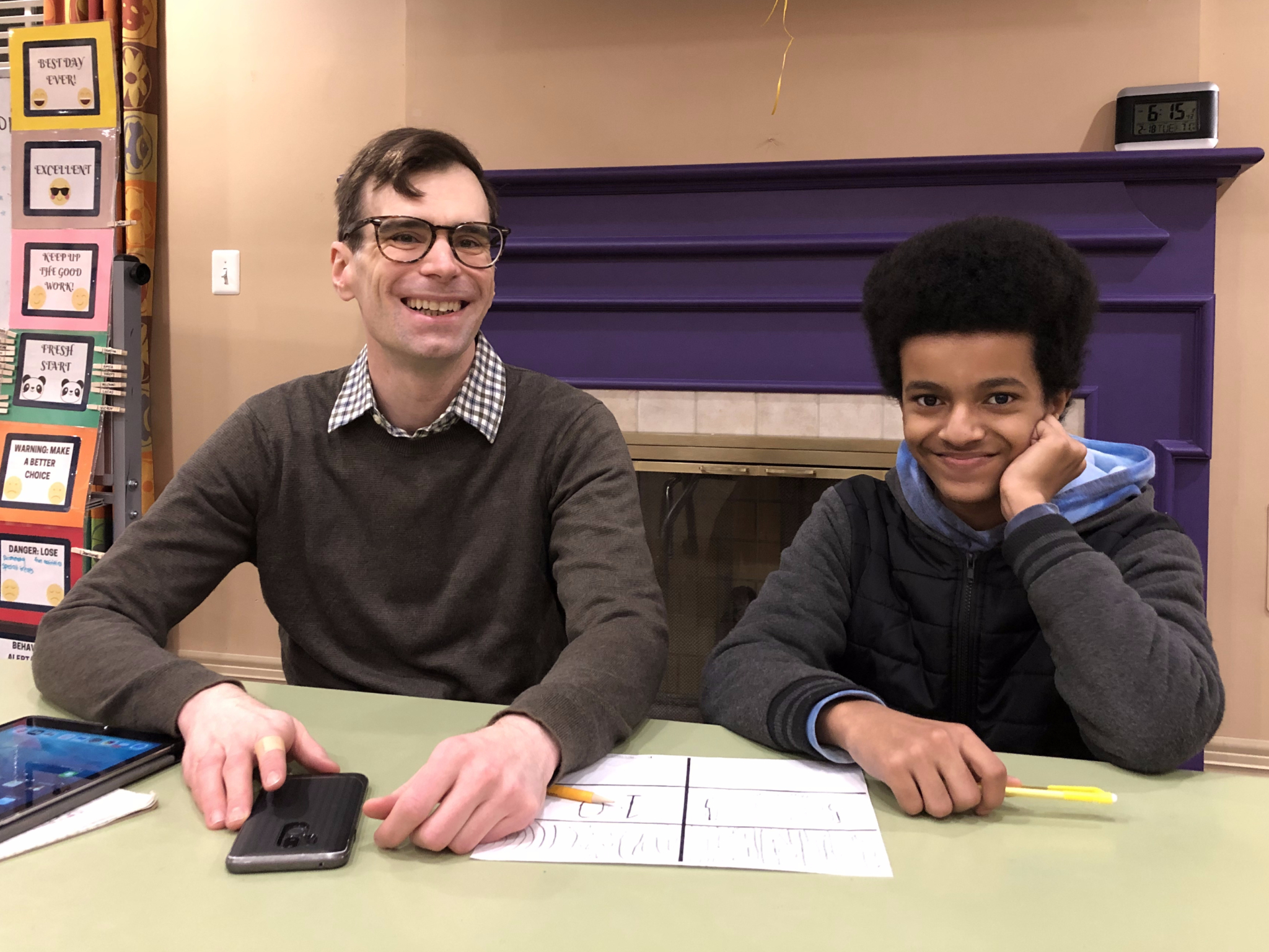 Two people sit at a table smiling. The adult on the left wears glasses and a brown sweater, while the young person on the right rests their head on their hand. Papers, a phone, and a highlighter are on the table.
