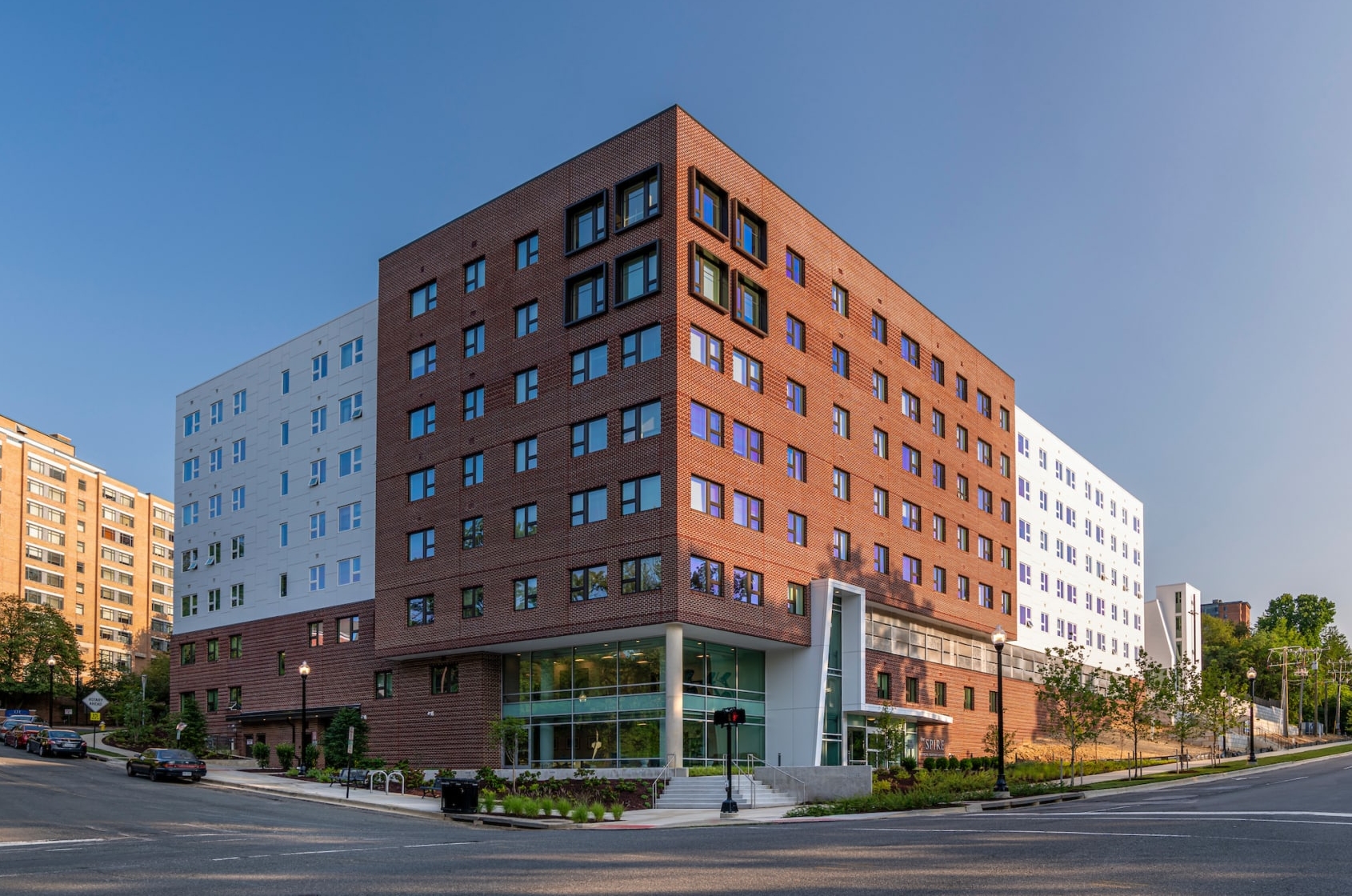 A modern mid-rise apartment building with red and white facades stands at a street corner, surrounded by trees, sidewalks, and adjacent to other urban buildings under a clear blue sky.