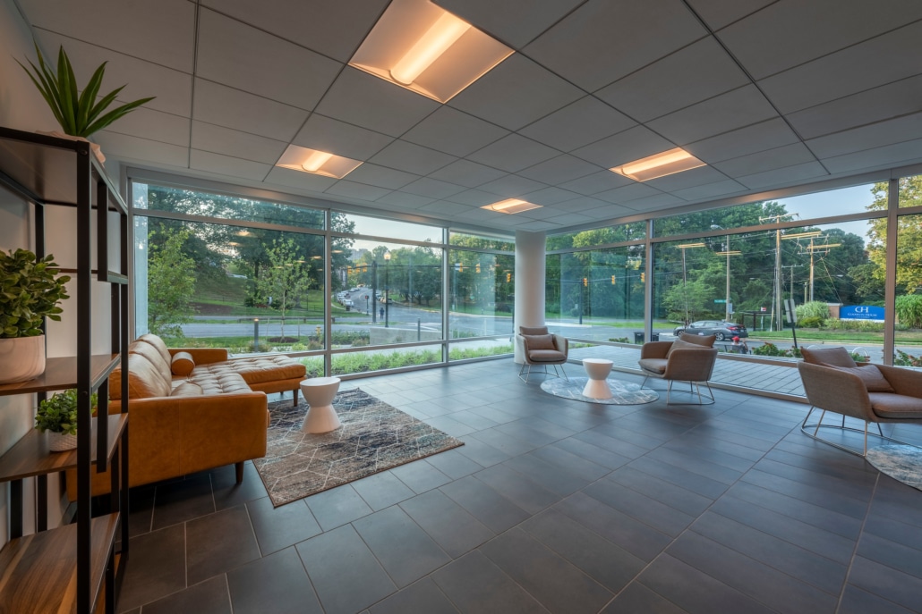 Modern office lobby with large windows, tan sofas, gray armchairs, glass tables, potted plants, a gray tile floor, and natural light streaming in, overlooking a street and green trees outside.