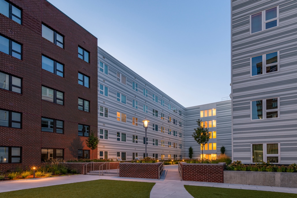 A modern apartment complex features a courtyard with grass, small trees, and raised brick planters. The buildings have brick and striped facades with many windows, and some apartment lights are on at dusk.