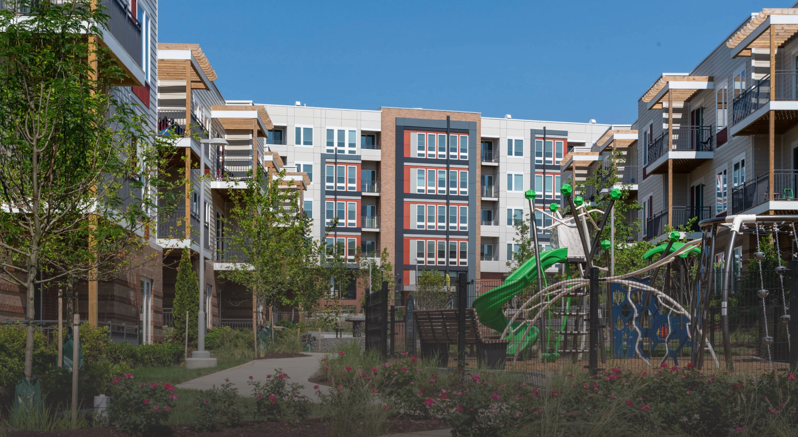 A modern apartment complex with multiple stories and large windows surrounds a courtyard featuring a playground, benches, and vibrant greenery—an inviting space within affordable homes & communities under a clear blue sky.