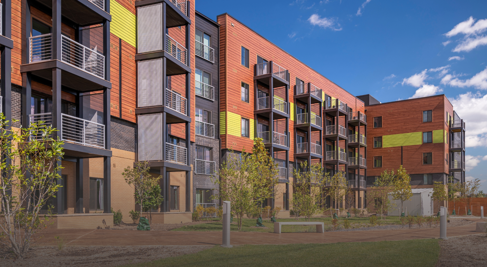 Modern multi-story apartment building with wood and metal exterior, balconies, and yellow accents. Affordable Homes & Communities features landscaped greenery, a paved walkway, and benches in the courtyard under a partly cloudy blue sky.