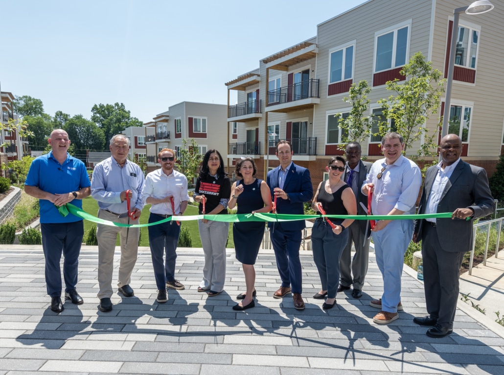 A group of people stand in front of new apartment buildings, holding a large green ribbon and oversized scissors, celebrating a ribbon-cutting ceremony on a sunny day.