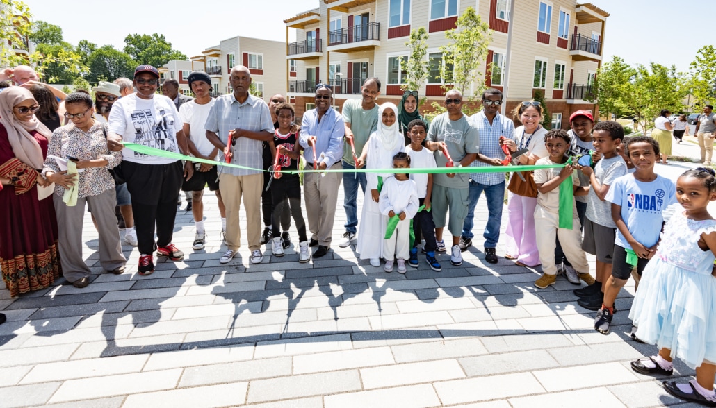 A diverse group of adults and children smile and pose at an outdoor ribbon-cutting ceremony in front of modern apartment buildings on a sunny day.